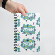 Load image into Gallery viewer, A spiral notebook being held by a person. The white spiral notebook has black spiral binding. The pattern design features the word &quot;dream&quot; inside a cloud shape with sketchy elements around. 