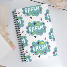 Load image into Gallery viewer, A spiral notebook on a white, wood tabletop. The white spiral notebook has black spiral binding. The pattern design features the word &quot;dream&quot; inside a cloud shape with sketchy elements around. 