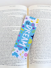 Load image into Gallery viewer, A bookmark placed inside a book. The bookmark design has the word &quot;dream&quot; inside a teal cloud-like shape with sketchy, whimsical items around. 