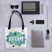 Load image into Gallery viewer, A white tote bag with black handles on a grey tabletop with a laptop and other office supplies. The bag features a design with the word &#39;dream&#39; with an illustrated cloud around the word with colorful sketchy around. 