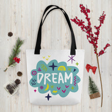 Load image into Gallery viewer, A white tote bag with black handles on a wood tabletop with Christmas items around it. The bag features a design with the word &#39;dream&#39; with an illustrated cloud around the word with colorful sketchy around. 