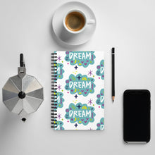 Load image into Gallery viewer, A spiral notebook on a tabletop with a coffee mug and phone around it. The white spiral notebook has black spiral binding. The pattern design features the word &quot;dream&quot; inside a cloud shape with sketchy elements around. 