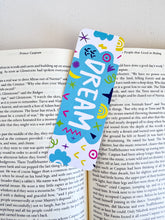 Load image into Gallery viewer, A bookmark placed inside a book. The bookmark design has the word &quot;dream&quot; inside a teal cloud-like shape with sketchy, whimsical items around. 