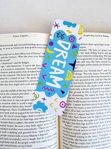 A bookmark placed inside a book. The bookmark design has the word "dream" inside a teal cloud-like shape with sketchy, whimsical items around. 