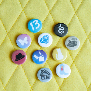 A photo of 10 pin badges on a yellow background. The badges feature illustrations of a snake, a lighter, a cowboy boot,  cat, a butterfly, a house, a typewriter, a diamond, the number 13, and a black hat. 