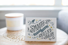 Load image into Gallery viewer, A greeting card laying on a wooden table with some cut wood details. The card features the words “A smooth sea never made a skilled sailor.”