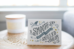 A greeting card laying on a wooden table with some cut wood details. The card features the words “A smooth sea never made a skilled sailor.”