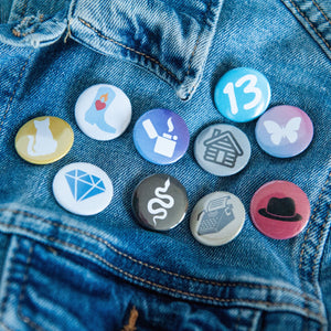 A photo of 10 pin badges on a jean jacket. The badges feature illustrations of a snake, a lighter, a cowboy boot,  cat, a butterfly, a house, a typewriter, a diamond, the number 13, and a black hat. 