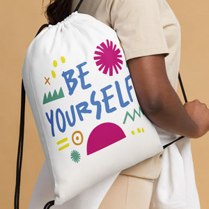 A woman holding a white drawstring bag over her shoulder. The drawstring bag has the words "Be Yourself" in blue with green illustrated sketchy pieces around the words. 