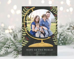 A photo of a one-sided Christmas card showing the front of the card standing up with pine needles behind and blurred white Christmas lights. The photo card features a photo inside an illustrated globe. Around the globe are illustrated leaves. At the bottom of the globe the words “Hope of the World.”