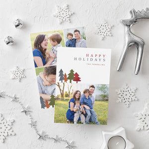 A photo of a double-sided Christmas card showing the front and back of the card laying on a white surface. Around the two sides of the card are surrounded with white snowflake ornaments, silver bells, a silver reindeer, white tea light snowflake candle and silver star garland. The front of the card features a photo on the bottom and on the top it reads “Happy Holidays, The Franklins” with illustrated modern pine trees. The back of the card features three photos. 