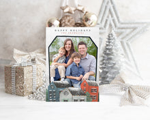 Load image into Gallery viewer, A photo of a one-sided Christmas card showing the front of the card standing up with Christmas items behind the card. There’s a sparkly star, glitter wrapped gift, ribbon and ornaments with a small silver tree around the photo card. The photo card features the words “Happy Holidays, The Franklins” at the top with a photo featured in a house shaped frame. Below the photo are illustrated houses covered in snow. 