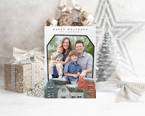 A photo of a one-sided Christmas card showing the front of the card standing up with Christmas items behind the card. There’s a sparkly star, glitter wrapped gift, ribbon and ornaments with a small silver tree around the photo card. The photo card features the words “Happy Holidays, The Franklins” at the top with a photo featured in a house shaped frame. Below the photo are illustrated houses covered in snow. 