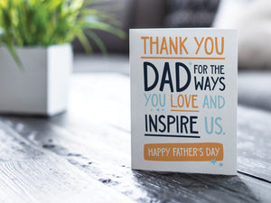 A greeting card is featured on a desktop with a green plant to the side. The card features the words  "Thank You Dad for the ways you love and inspire us. Happy Father's Day.” 