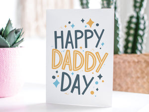 A greeting card featured standing up on a white tabletop with a pink plant pot in the background and some succulents in the pot. There’s a woven basket in the background with a cactus inside. The card features the words “Happy Daddy Day” with diamond shapes surrounding the letters. 