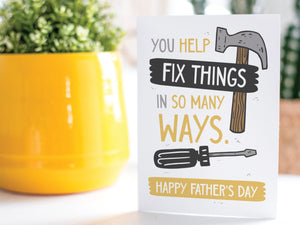 A greeting card is on a table top with a yellow plant pot and a green plant inside. The card features the words “You Help Fix Things in so Many Ways, Happy Father's Day” with an illustrated hammer and screwdriver around the words. 