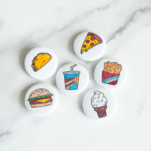 A set of six button pins on a white tabletop. The pins feature illustrations of a taco, pizza slice, french fries, an ice cream cone, a milkshake and a burger. 