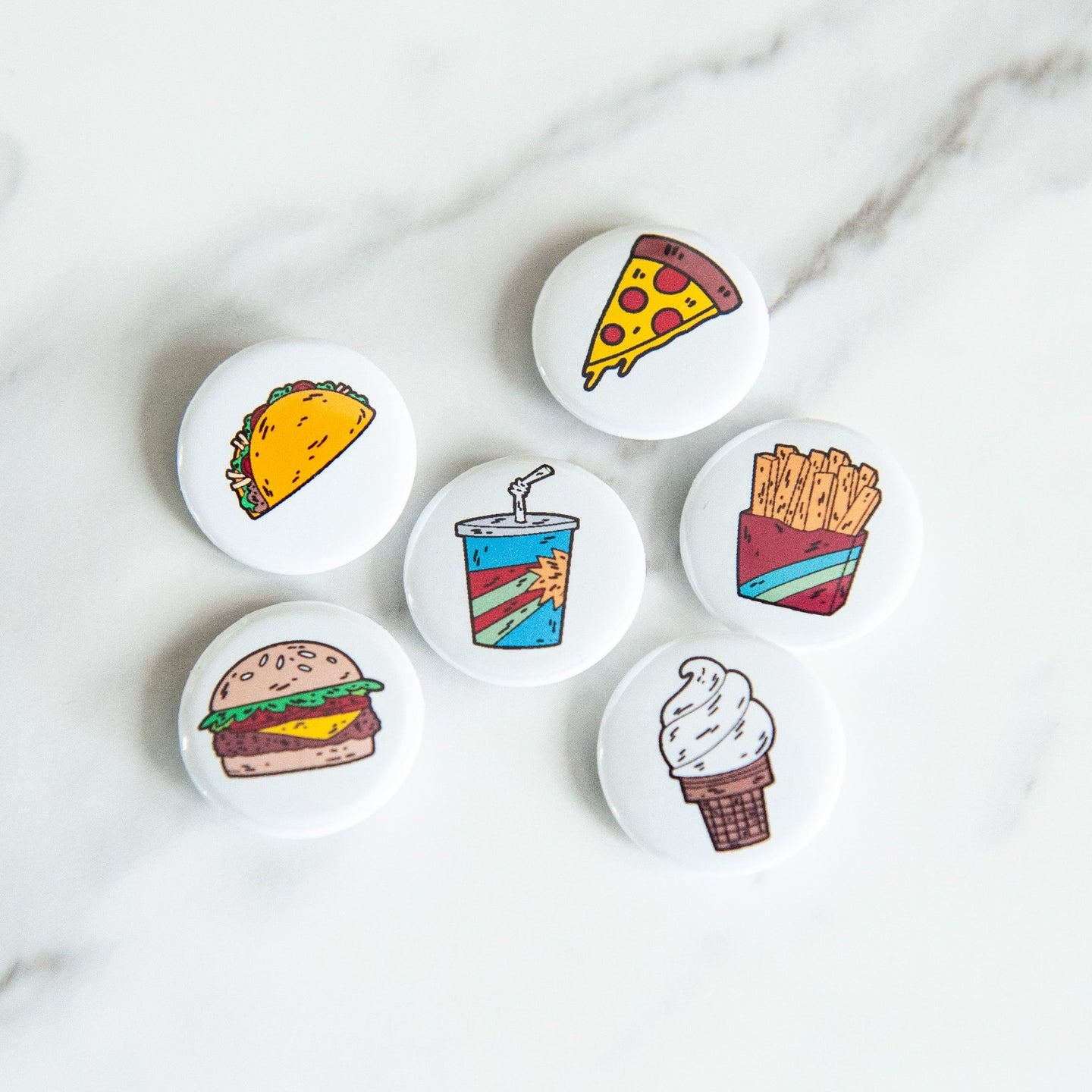 A set of six button pins on a white tabletop. The pins feature illustrations of a taco, pizza slice, french fries, an ice cream cone, a milkshake and a burger. 