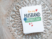 Load image into Gallery viewer, A greeting card laying on a wooden table with some cut wood details. The card features the words “For my Husband on Father&#39;s Day” with small hearts around it. 