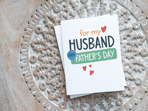 A greeting card laying on a wooden table with some cut wood details. The card features the words “For my Husband on Father's Day” with small hearts around it. 