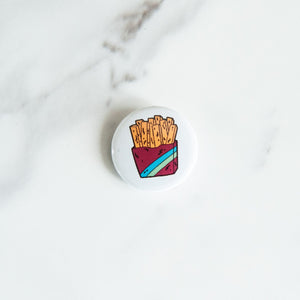 A button pin on a white tabletop. The pin features an illustration of french fries.