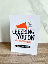 Load image into Gallery viewer, A card sitting on a tabletop. The card features the words “Cheering You on into This New Year, Happy Birthday.&quot;