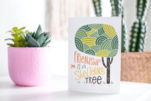 Load image into Gallery viewer, A greeting card featured standing up on a white tabletop with a pink plant pot in the background and some succulents in the pot. There’s a woven basket in the background with a cactus inside. The card features the words “Friendship is a sheltering tree” with an illustrated tree.