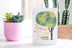 A greeting card featured standing up on a white tabletop with a pink plant pot in the background and some succulents in the pot. There’s a woven basket in the background with a cactus inside. The card features the words “Friendship is a sheltering tree” with an illustrated tree.