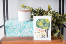 Load image into Gallery viewer, A greeting card is on a table top with a present in blue wrapping paper in the background. On top of the present is a candle and some greenery from a plant too. The card features the words “Friendship is a sheltering tree” with an illustrated tree. 