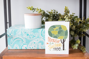 A greeting card is on a table top with a present in blue wrapping paper in the background. On top of the present is a candle and some greenery from a plant too. The card features the words “Friendship is a sheltering tree” with an illustrated tree. 