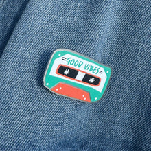 Load image into Gallery viewer, An enamel pin featuring the words &quot;good vibes&quot; inside an illustrated cassette tape. The pin is on a jean jacket.