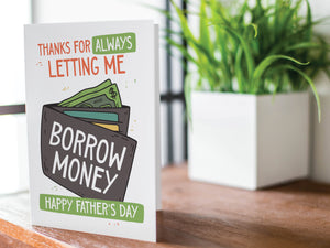 A greeting card is featured on a wood coffee table with a green plant in a white planter in the background. The card features the words "Thanks for Always Letting Me Borrow Money, Happy Father’s Day.” 