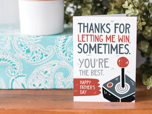 A greeting card is on a table top with a present in blue wrapping paper in the background. On top of the present is a candle and some greenery from a plant too. The card features the words  "Thanks for letting me win, sometimes. You're the best. Happy Father's Day." 