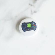 Load image into Gallery viewer, A button pin on a white tabletop. The button features an illustration of a retro game controller. 