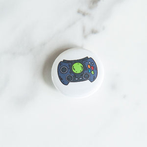 A button pin on a white tabletop. The button features an illustration of a retro game controller. 