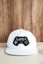 Load image into Gallery viewer, A white trucker hat on a tabletop. The hat features an embroidered design with an illustrated game controller with the word &#39;gamer&#39; inside the illustration.  