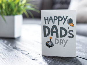 A greeting card featured on a black, wood coffee table. There’s a white planter in the background with a green plant. There’s also a gray sofa in the background with a white pillow. The card features the words “Happy Dad’s Day” with an illustrated game controller and hat. 