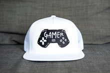 Load image into Gallery viewer, A white trucker hat sitting on a gray sofa. The hat features an embroidered design with an illustrated game controller with the word &#39;gamer&#39; inside the illustration.  