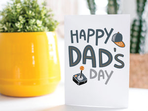A greeting card is on a table top with a yellow plant pot and a green plant inside. The card features the words “Happy Dad’s Day” with an illustrated game controller and hat. 