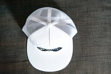 Load image into Gallery viewer, A white trucker hat on a gray sofa showing the top of the hat. The hat features an embroidered design with an illustrated game controller with the word &#39;gamer&#39; inside the illustration.  