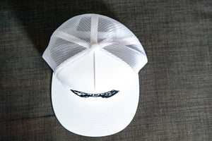 A white trucker hat on a gray sofa showing the top of the hat. The hat features an embroidered design with an illustrated game controller with the word 'gamer' inside the illustration.  