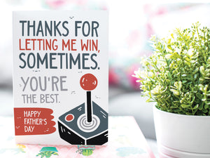 A greeting card is on a table top with a gift in pink wrapping paper. Next to the gift is a white plant pot with a green plant. The card features the words "Thanks for letting me win, sometimes. You're the best. Happy Father's Day." 