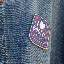 Load image into Gallery viewer, An enamel pin featured on a jean jacket. The pin has the words &#39;I love books&quot; with the word love shown as a heart. The words are featured inside an illustrated book, all in bright fun colors. 