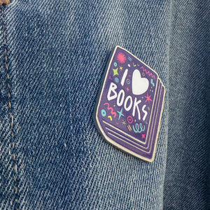 An enamel pin featured on a jean jacket. The pin has the words 'I love books" with the word love shown as a heart. The words are featured inside an illustrated book, all in bright fun colors. 