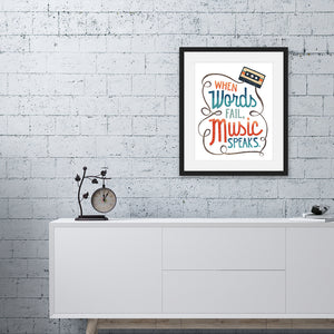 Artwork is featured in a black frame on a white, brick wall. The art is hanging above a white credenza. The artwork features hand drawn lettering with the phrase "When words fail, music speaks." In the upper corner of the words an illustrated cassette tape is featured.