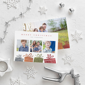 A photo of a double-sided Christmas card showing the front and back of the card laying on a white surface. Around the two sides of the card are surrounded with Christmas items. The front of the card features three photos with illustrated gifts on the bottom. On top of the photos the words “Merry Christmas” is featured with a family name below which you can edit. The back of the card features one photo with some more illustrated gifts. 