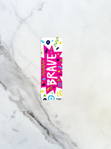 A bookmark with the word "brave" in white within a star-like pink shape with fun, sketchy elemts around. The bookmark is on a tabletop.