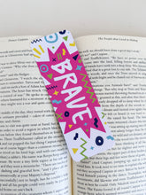 Load image into Gallery viewer, A bookmark with the word &quot;brave&quot; in white within a star-like pink shape with fun, sketchy elemts around. The bookmark is placed inside a book. 