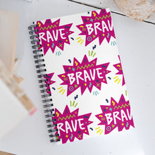 Load image into Gallery viewer, A spiral notebook on a white, wood tabletop. The white spiral notebook has black spiral binding. The pattern design features the word &quot;brave&quot; inside a hot pink star with sketchy elements around. 