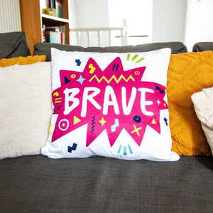 A white pillow on a grey sofa with other pillows. The white pillow features the artwork on a white background with a hot pink star with the word 'brave' inside and sketchy, fun elements around the word. 